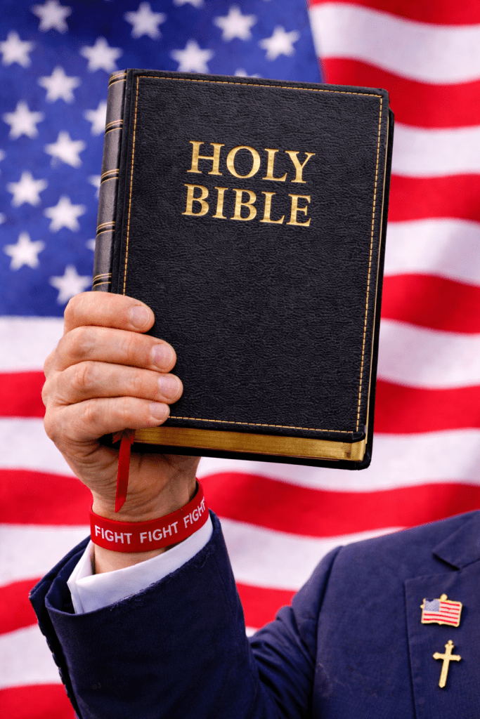 A person holding a black Holy Bible against a backdrop of the American flag, wearing a red rubber bracelet that reads 'FIGHT'.